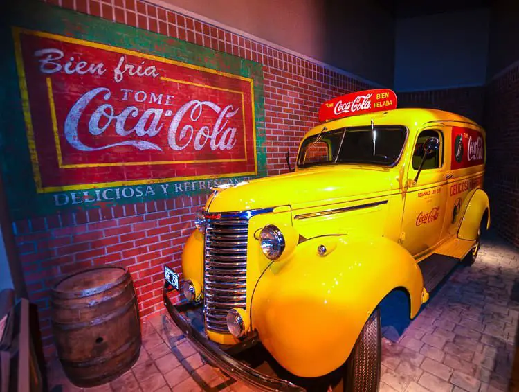A vintage yellow delivery truck with the Coca-Cola logo parked in front of a brick wall adorned with a nostalgic Coca-Cola advertisement.