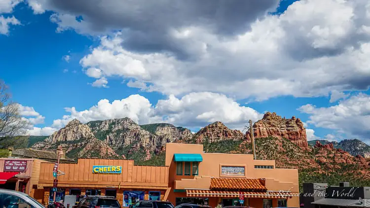 Downtown Sedona bustling with activity, with shops and a sign that says 'CHEERS' in the foreground and majestic red rock formations under a partly cloudy sky in the background.