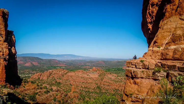 A panoramic view of Sedona's red rock formations with a clear blue sky. A person stands at the edge of a cliff, emphasizing the vastness of the landscape.