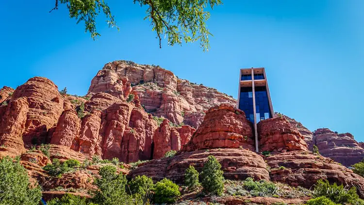 The Chapel of the Holy Cross, a modern architectural marvel built into the red rocks of Sedona, with clear blue skies above and a foreground of lush green trees.