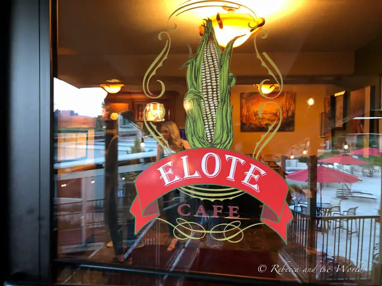 The glass door of Elote Cafe, reflecting the interior and exterior seating areas, with the restaurant's distinctive corn-inspired logo displayed prominently.