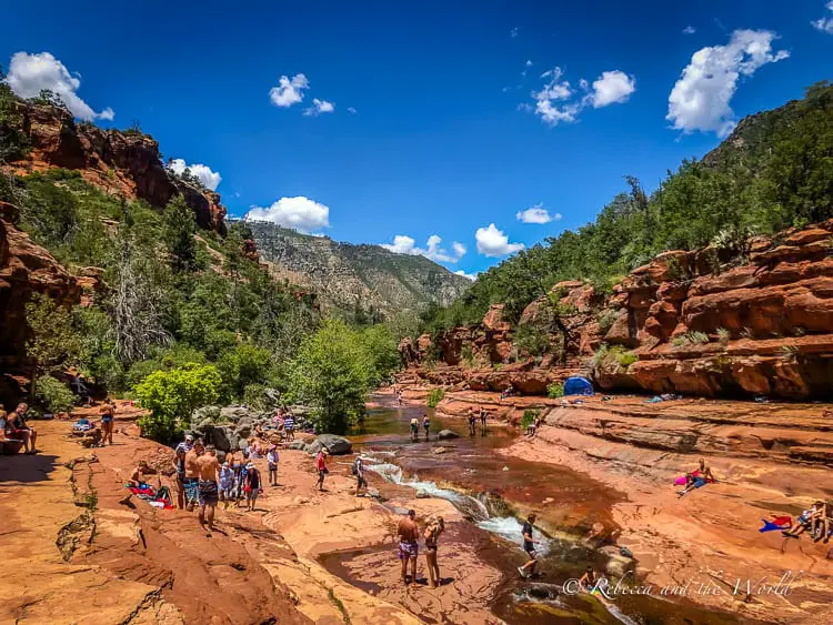 A vibrant scene at the popular Slide Rock State Park in Sedona where people are enjoying the water, surrounded by towering red rock formations and greenery under a blue sky.