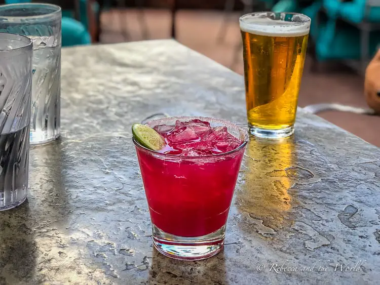 A close-up of a vibrant red cocktail - a prickly pear margarita - with ice and a lime wedge, beside a pint glass of golden beer, both resting on a textured table surface, embodying the casual dining experiences in Sedona.