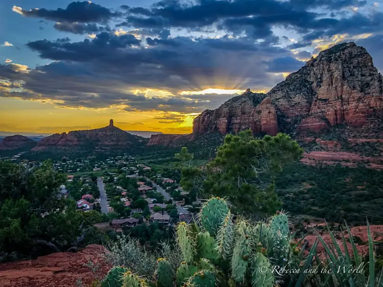 A scenic sunset view over Sedona, showcasing layered red rock formations and the silhouette of a distinctive rock spire, with residential areas nestled among the verdant valley.