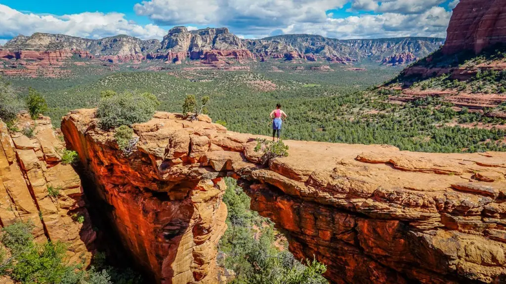 A breathtaking perspective from atop a natural sandstone bridge in Sedona, with lush greenery and rugged terrain extending into the distance. This is the view from the top of the Devil's Bridge Trail.