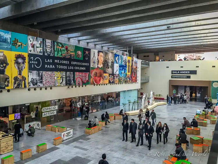 A spacious interior of the modern Centro Cultural La Moneda in Santiago Chile with people walking about. Above them is a gallery of colourful banners featuring various artistic styles. Visiting an exhibition at the centre is one of the best things to do in Santiago.