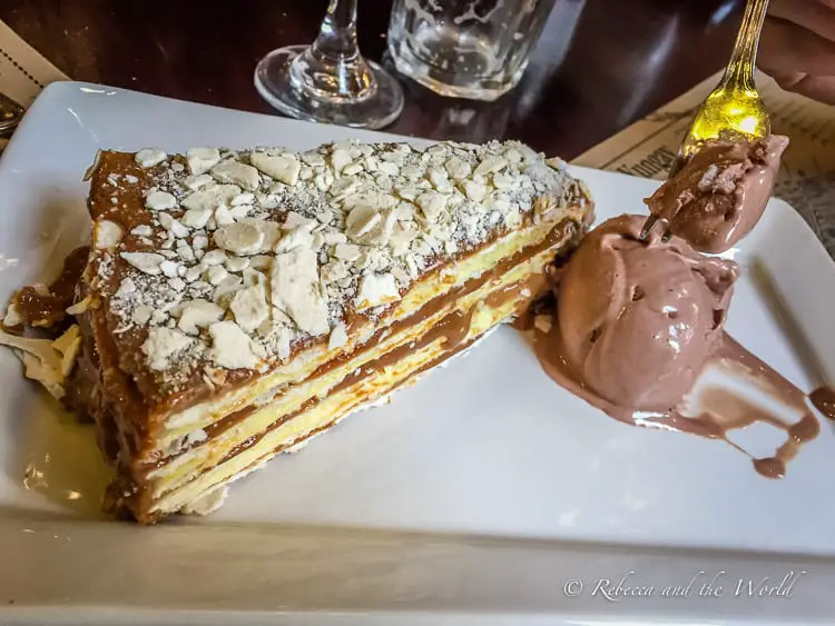 A plated dessert consisting of a layered pastry with a dusting of powdered sugar and almond flakes on top, accompanied by a scoop of chocolate ice cream with a golden spoon partially submerged in it. The table shows two empty glasses in the background, suggesting a recently enjoyed meal.