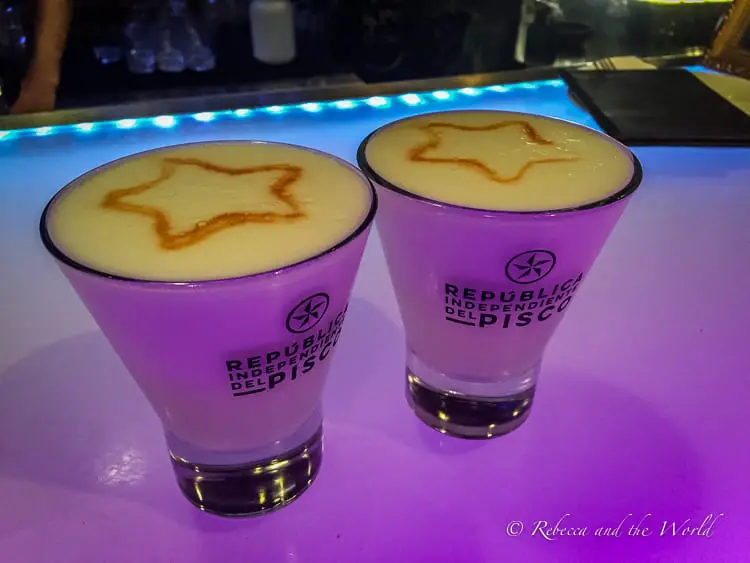 Two glasses of Pisco Sour on a bar counter with frothy tops and caramel designs resembling stars, illuminated by purple-blue light.