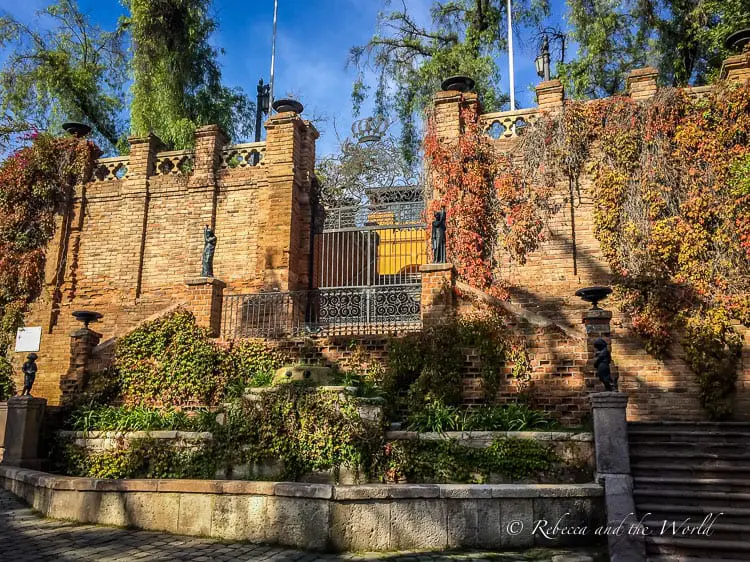 A brick structure with ornate metal gates, overgrown with ivy and other plants, in a garden setting. This is Cerro Santa Lucia, one of the best places to visit in Santiago Chile.