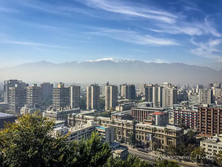 A view of Santiago Chile with numerous high-rise buildings under a blue sky. In the background, the Andes mountain range with snow-capped peaks is partially obscured by haze.