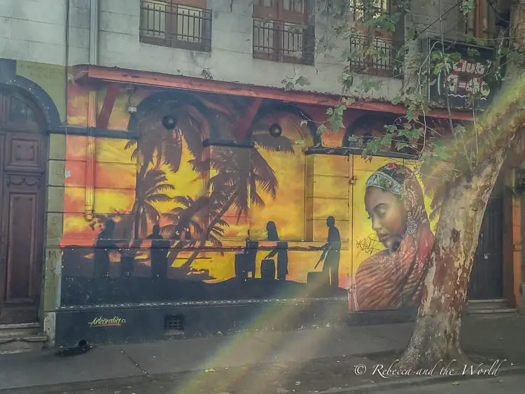 A large mural painted on a building's wall depicting a tropical scene with palm trees, silhouettes of people playing drums, and a woman's face in profile. Bellavista, a neighbourhood in Santiago Chile, is known for its street art and is one of the best things to see in Santiago.