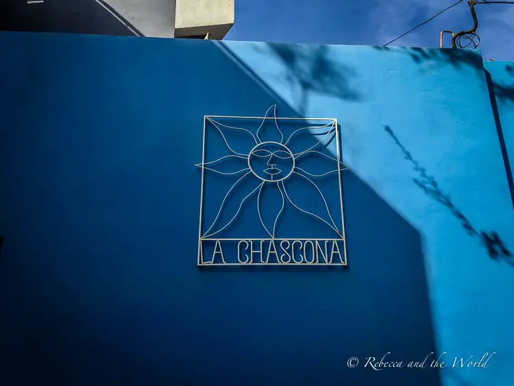 A metal sign featuring a stylized sun design and the text "LA CHASCONA" affixed to a deep blue wall, with shadows of branches cast on the wall.
