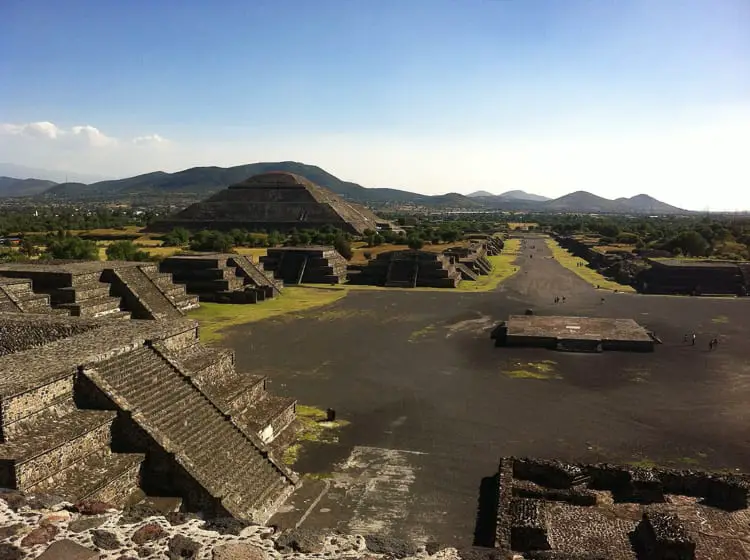 Aerial view of the ancient Mesoamerican city of Teotihuacan, showcasing the Avenue of the Dead and the Pyramid of the Sun, with a scenic mountain backdrop under a clear sky. Teotihuacan is an ancient site outside of Mexico City.