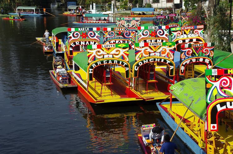 Colorful trajineras, traditional Mexican flat-bottomed boats, lined up along the canals of Xochimilco, adorned with vibrant decorations and names painted on the sides. Grab some friends and rent a boat to explore Xochimilco in Mexico City.