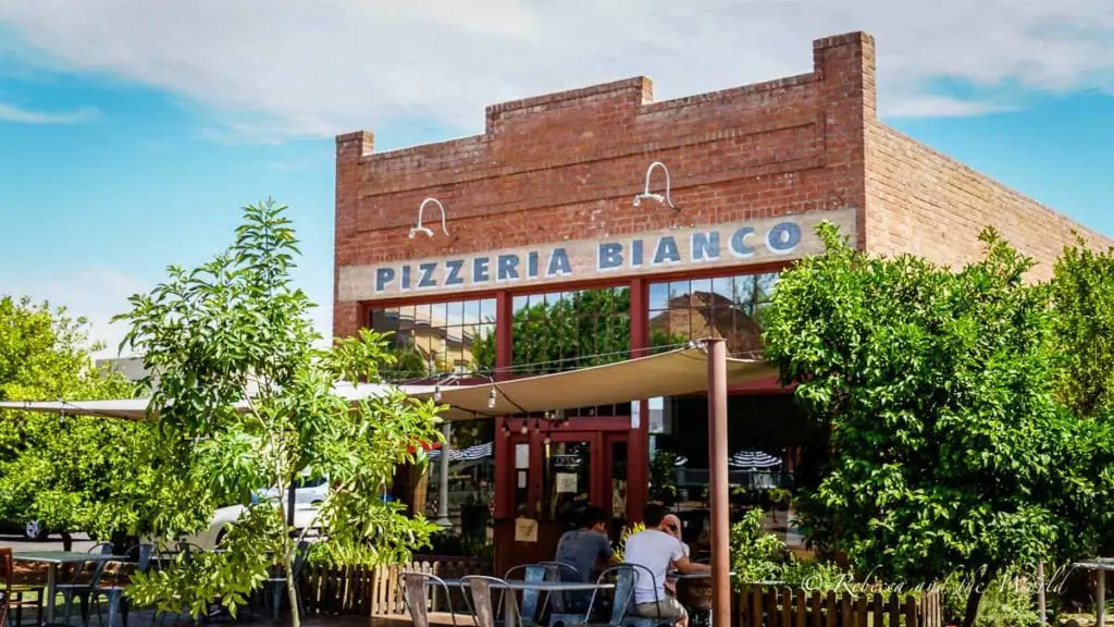 The front facade of Pizzeria Bianco, a brick building with a large sign over a covered patio where patrons are seated. Pizzeria Bianco in Phoenix AZ serves up some of the best pizza in the entire country.