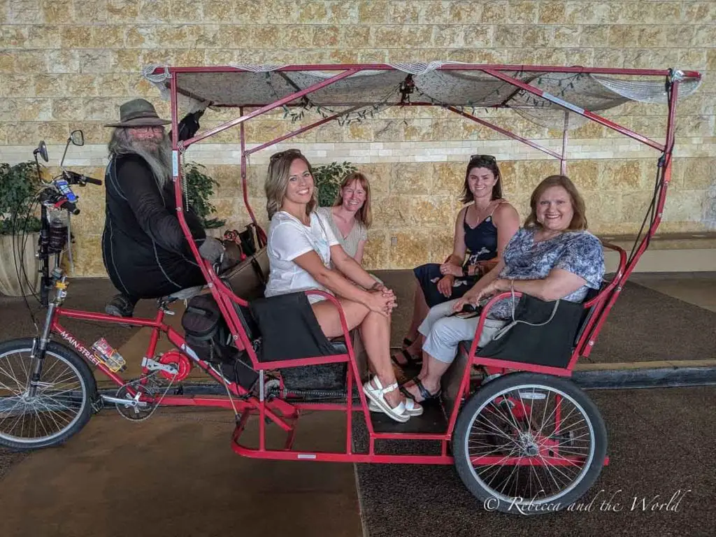 Four women and a bearded cyclist with a hat sitting on a red pedicab, smiling at the camera. Take a pedicab tour of Phoenix's murals while you have 36 hours in Phoenix.