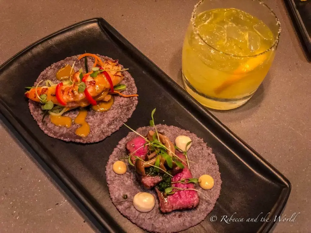 A dish with two purple corn tortillas topped with grilled seafood and sliced vegetables, accompanied by a glass of a yellow, citrusy beverage with ice. The tacos at CRUjientes are so delicious - try to go during happy hour on your visit to Phoenix.