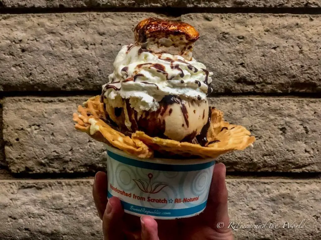 A hand holding a waffle cone with a scoop of ice cream topped with whipped cream and chocolate sauce against a brick wall. The ice cream at Sweet Republic is divine - a must-eat when you're in Phoenix for 36 hours!