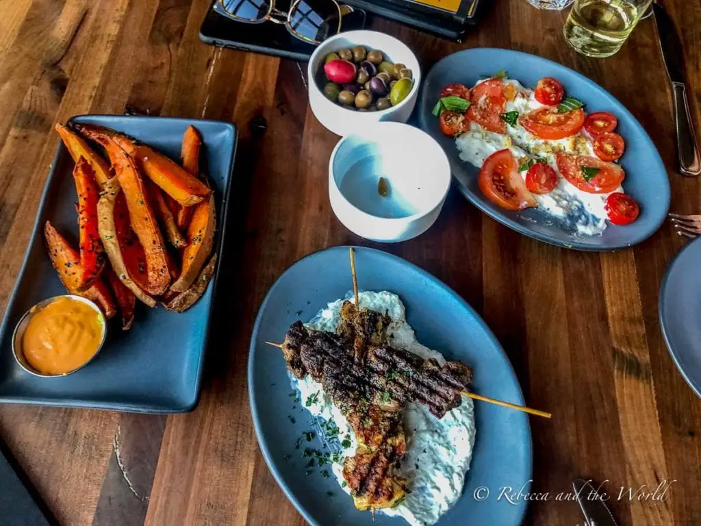 A table with dishes of food including sweet potato fries, skewered meat on a creamy white sauce, and a tomato and cheese salad, with a glass of white wine. Grab dinner at Postino when you visit Phoenix.