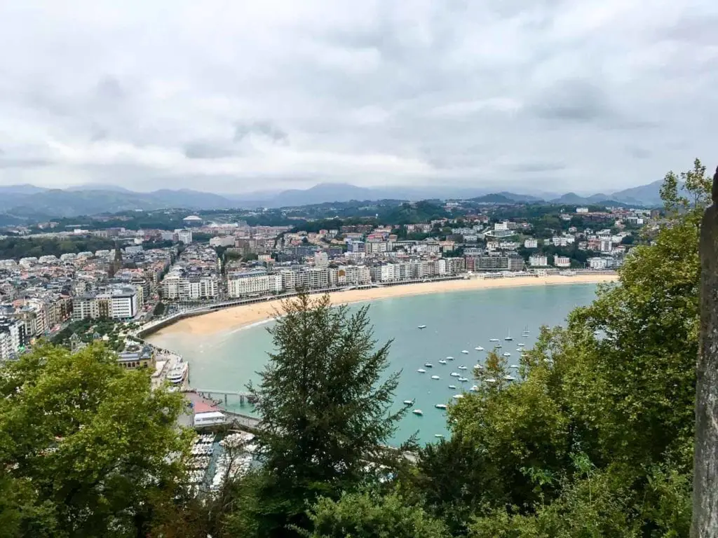Overlooking La Concha Beach from a high vantage point, the image captures the sweep of the sandy shore and the turquoise sea. The urban landscape of San Sebastian stretches into the rolling hills in the background, under a cloudy sky. Monte Urgull has stunning views over San Sebastian.