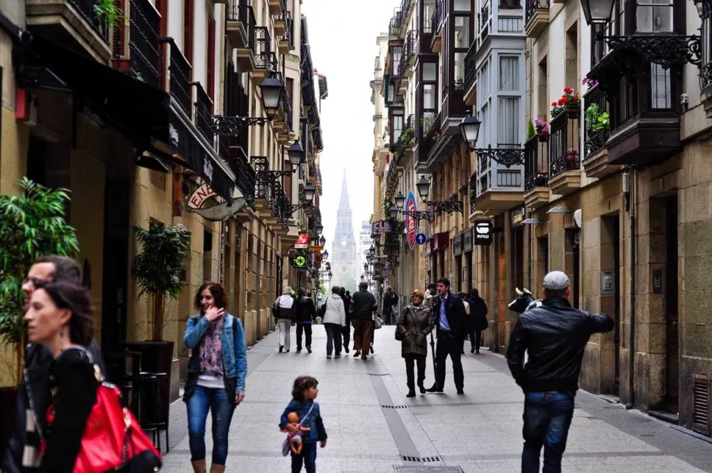 A bustling street scene in San Sebastian with people walking along a narrow lane flanked by traditional buildings. A pointed church spire rises in the distance, drawing the eye along the street's perspective. Getting lost in the Old Town is one of the best things to do in San Sebastian.