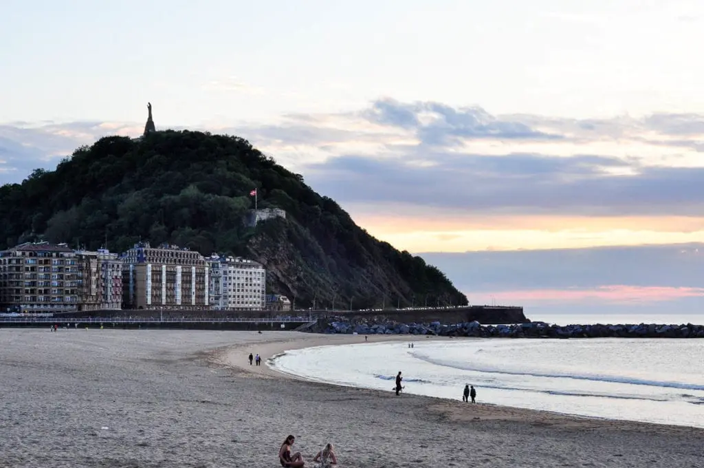 Dusk at San Sebastian's beach, showing the silhouette of Mount Urgull with its prominent statue and flag. The beachfront is lined with buildings, and people are visible enjoying the shore as the sky fades into sunset colours. One of the most popular things to do in San Sebastian is relax on Playa de la Concha, or Shell Beach.