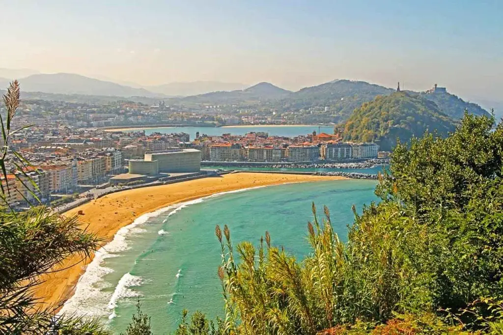 A bright daytime view of San Sebastian, with La Concha Beach in the foreground and the city's architecture behind. The lush greenery of Mount Urgull adds contrast to the urban landscape under a clear blue sky. There are also great hiking trails around San Sebastian in northern Spain.