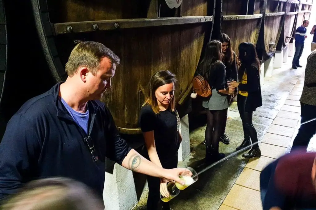 An individual practicing the traditional Basque method of pouring cider from a height to aerate it, with onlookers in a rustic cidery, exemplifying the region's rich cider culture. Only available seasonally, visiting a Basque cider house to taste the local cider paired with an amazing feast is an incredible thing to do in San Sebastian.