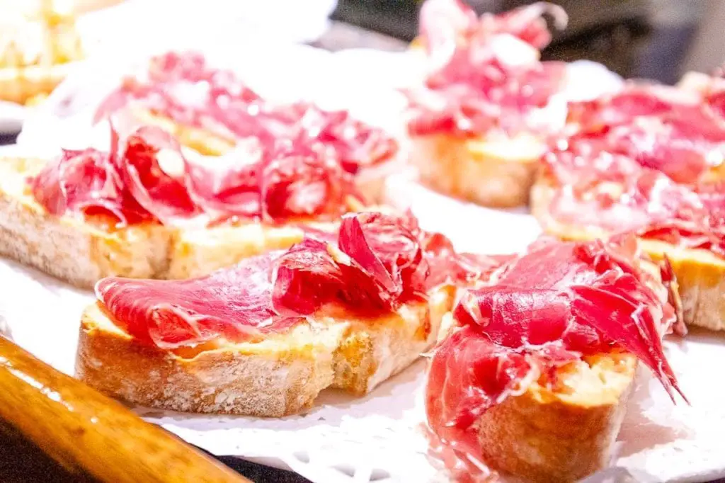 A selection of pintxos featuring Iberico ham on slices of crusty bread, displayed prominently on a bar counter, indicative of the culinary delights found in San Sebastian. After trying pintxos at the local bars, try your hand at making them yourself.