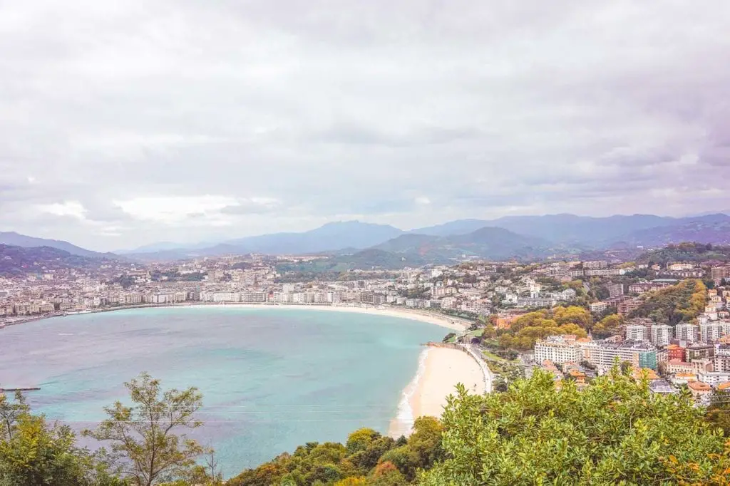A panoramic view of San Sebastian's bay from a high perspective, showing the curve of La Concha Beach, the cityscape, and the mountains in the distance, all under a pastel-toned sky. Monte Igueldo in San Sebastian offers stunning views over the city.