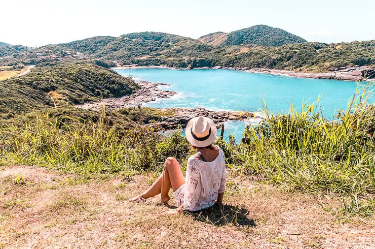A woman in a straw hat sitting on a grassy hill overlooking the turquoise waters of a secluded bay in Brazil's coastal region. Buzios is a paradisiacal island only a short trip from Rio de Janeiro.