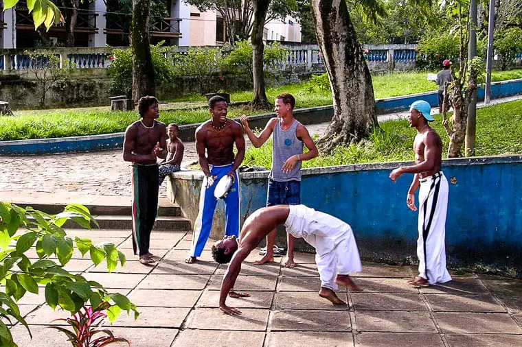 A group of young men practicing Capoeira, a Brazilian martial art that combines dance, acrobatics, and music, in a park setting, with one man performing an inverted kick while others watch and play instruments. Salvador in Brazil is the perfect mix of beaches, history, architecture and wildlife.