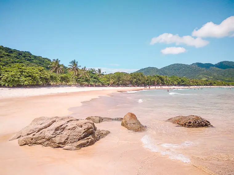 Pristine sandy beach lined with palm trees and backed by forested hills under a clear blue sky in Ilha Grande, Brazil. In Ilha Grande, you'll find some of Brazil's most beautiful beaches. Easily accessible from Rio de Janeiro, Ilha Grande is one of the most popular places to visit in Brazil.