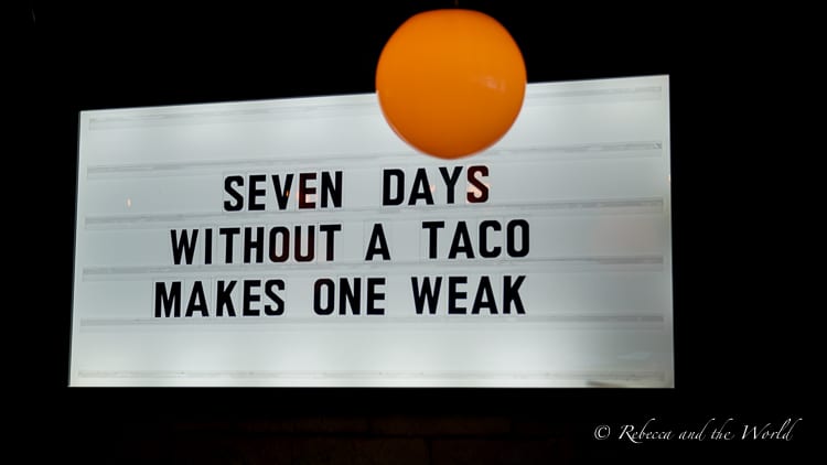 A humorous marquee sign that reads "Seven days without a taco makes one weak," set against a dark backdrop with a single orange balloon.