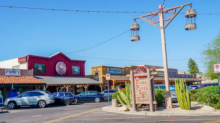 A bustling street view in Scottsdale showcasing an array of quaint shops and restaurants, with red wooden facades and a rustic charm, under a clear blue sky adorned with string lights.