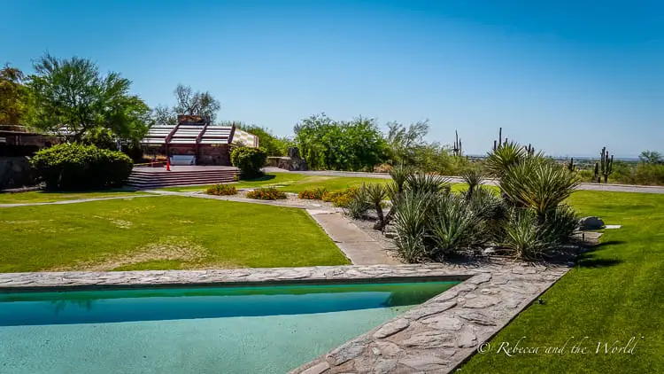 A well-manicured outdoor setting with a rectangular swimming pool, green lawns, and desert landscaping, featuring a view of a distinctive red-roofed building with large windows. This is Taliesen West, one of Frank Lloyd Wright's stunning buildings.