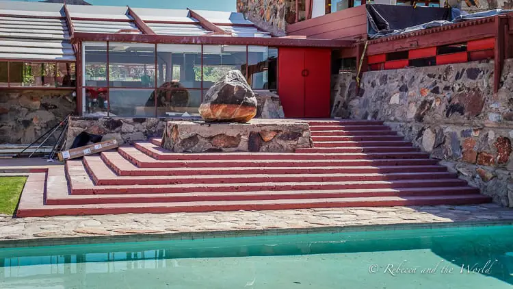 A red-roofed building, seen from across the swimming pool, highlighting the stone steps leading up to the glass doors and a striking boulder at the entrance. This is Taliesen West, one of Frank Lloyd Wright's stunning buildings.