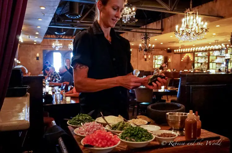 An attentive waitress preparing tableside guacamole in a warm, dimly-lit restaurant with a rustic aesthetic, complete with chandeliers and a full bar in the background.