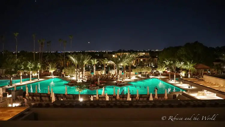 A night view of a resort pool illuminated with teal lights, with the glow of the pool lighting up the surrounding palm trees and loungers, creating a serene and inviting outdoor evening setting. This is one of the pools at The Phoenician hotel in Scottsdale, Arizona.