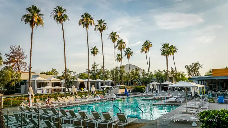 A daytime view of a resort pool lined with lounge chairs and tall palm trees, with mountains in the backdrop, under a partly cloudy sky, reflecting a relaxed and luxurious atmosphere. This is the amazing pool area at Andaz Scottsdale, one of the top hotels in Scottsdale.