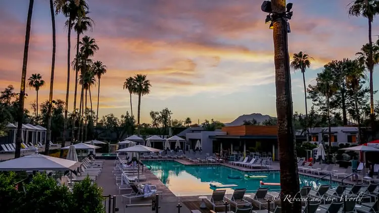 A luxurious poolside view at sunset with loungers and cabanas set against a backdrop of palm trees and a colorful sky reflecting the fading hues of the sun behind a mountain silhouette. This is the pool at the Andaz Scottsdale hotel.