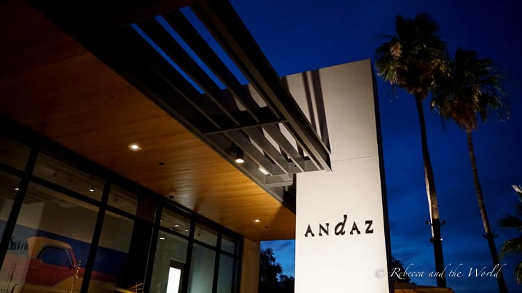 The entrance of the Andaz hotel in Scottsdale, Arizona, at dusk with modern architectural design, featuring sharp lines, wooden overhangs, and a sign bearing the hotel's name, flanked by tall palm trees against the twilight sky.