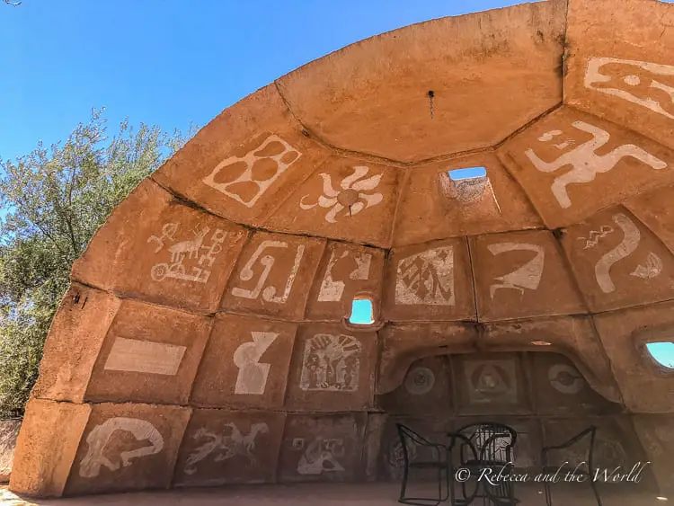 A semi-circular adobe structure with petroglyph-style engravings depicting various anthropomorphic and zoomorphic figures, with small openings allowing light to pass through. This is Cosanti, one of the most interesting things to do in Scottsdale, Arizona.