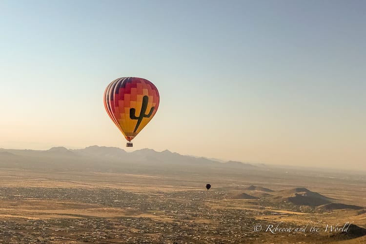 A serene early morning view of a hot air balloon adorned with a cactus design floating in the sky, with a landscape of mountains and the rising sun in the background. One of the best things to do on a weekend in Scottsdale is an early-morning hot air balloon ride.