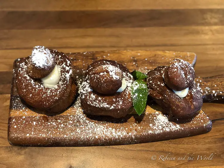 A dessert display of three spiral-shaped pastries filled with cream and dusted with powdered sugar, presented on a wooden cutting board garnished with fresh mint leaves.