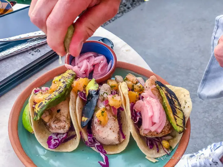 A close-up of a vibrant meal of fish tacos with colorful toppings like purple cabbage, mango salsa, and grilled avocado, served on a ceramic plate with a side of pickled onions. A hand is squeezing lime over the tacos.