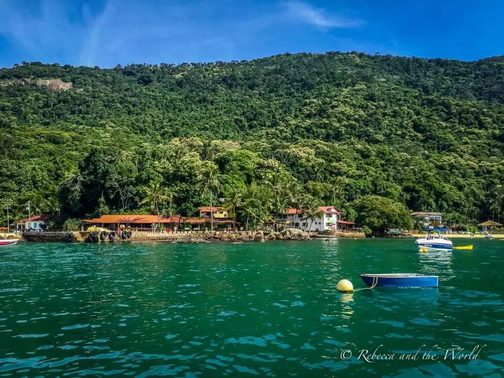 View of small houses and boats along the lush, green coastline of Ilha Grande, Brazil, with dense forest hills in the background. The first view you'll get of Ilha Grande, Brazil: lush green mountains.