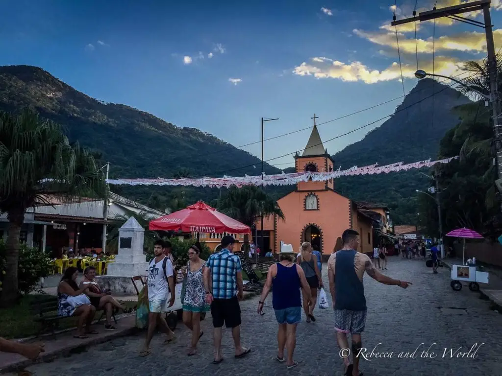 Vibrant street scene on Ilha Grande with people walking near a small church and local shops, under strings of lights and a mountain backdrop. The streets of Ilha Grande, Brazil, don't have cars.