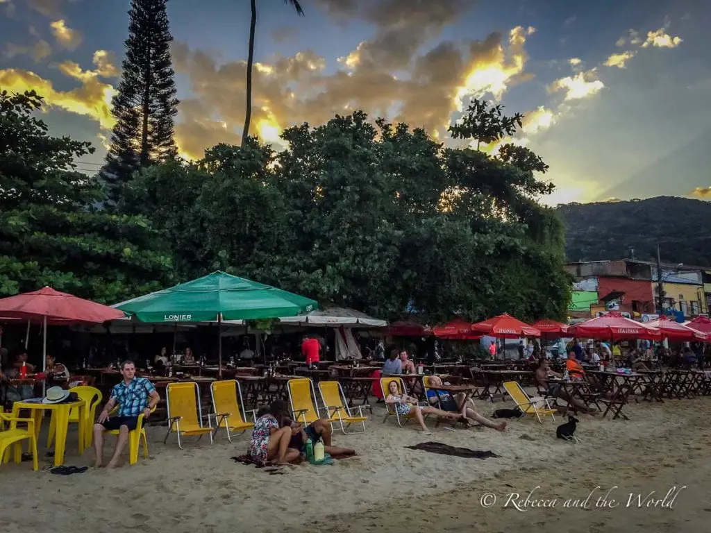 Busy beach scene at sunset on Ilha Grande, featuring numerous beach chairs and umbrellas, with people relaxing and enjoying the evening. Grab a seat on the beach in Ilha Grande and enjoy a caipirinha.