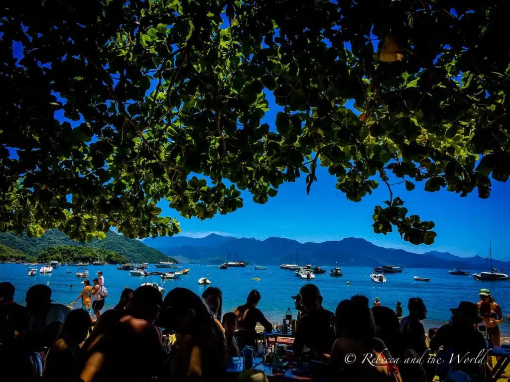 Under the shade of leafy branches, a view of Ilha Grande’s blue waters filled with boats, as seen from a busy beachside café. Enjoy the views while eating lunch or dinner at one of the restaurants in Ilha Grande.